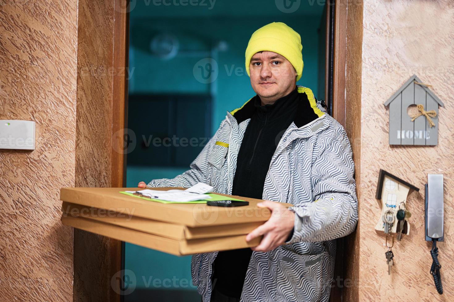 repartidor con cajas de cartón de pizza. mensajero con sombrero de color verde parado contra la puerta de una casa residencial foto