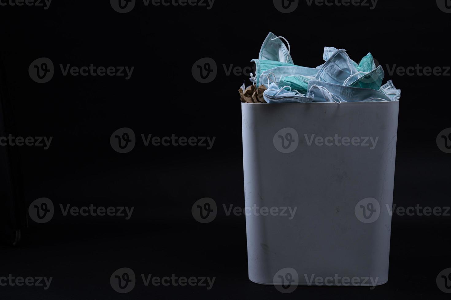Face mask in trash sanitary napkins, plastic. Medical waste unhygienic management concept. Disposal earloop face mask waste in garbage trash. Disgusting trash bin with medical waste backgound on black photo