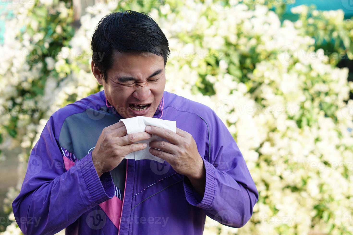 Asian man is sneezing, holding white tissue paper , open his mouth, standing beside blooming flowers at the park. Concept , Pollen allergy symptoms, sick, runny nose, catch a cold. health problems. photo