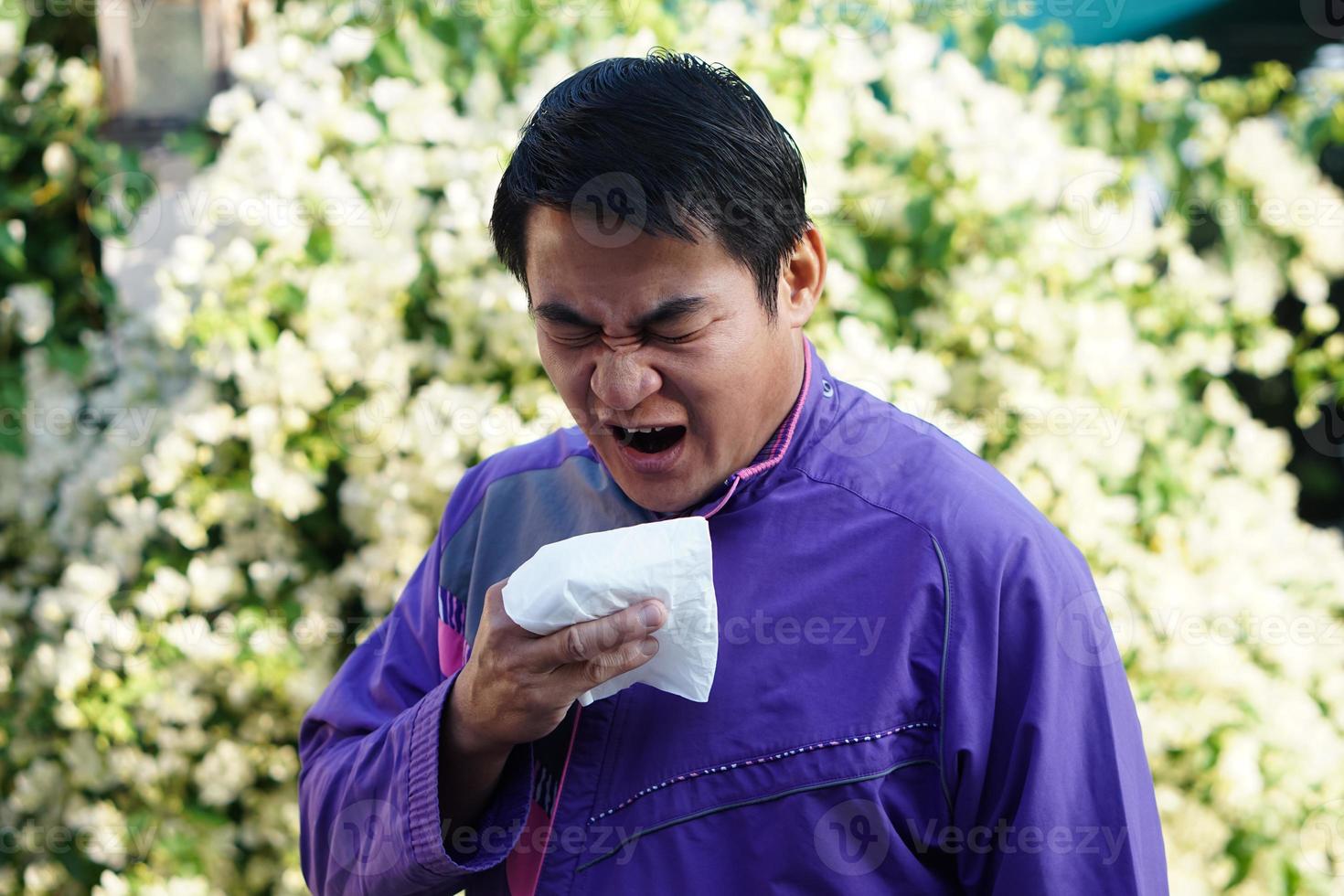 Asian man is sneezing, holding white tissue paper , open his mouth, standing beside blooming flowers at the park. Concept , Pollen allergy symptoms, sick, runny nose, catch a cold. health problems. photo