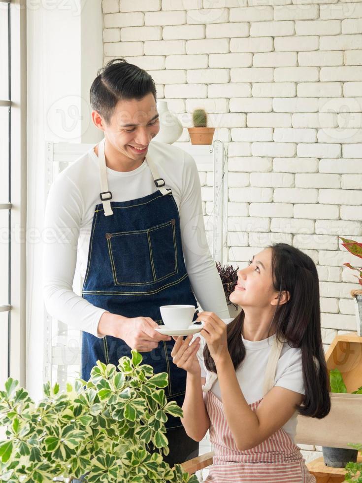 Portrait young boy girl Asian couple wearing apron. They are drinking hot coffee cup and helping to set up small plant on wooden table in their home. for beauty with bright smile  happy newlyweds photo
