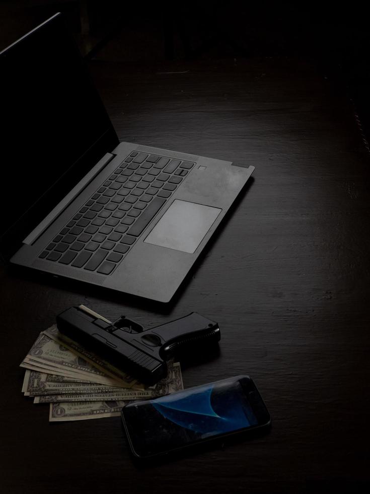 Computers, mobile phone, money, guns, and other devices that hackers use to steal or hijack information to trick victims into taking money. laying on wooden table in dark room in mysterious apartment. photo
