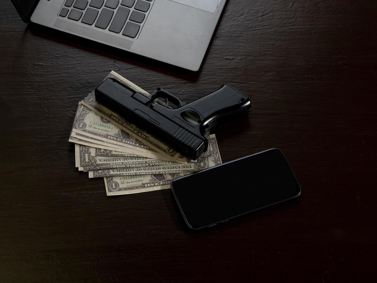 Computers, mobile phone, money, guns, and other devices that hackers use to steal or hijack information to trick victims into taking money. laying on wooden table in dark room in mysterious apartment. photo