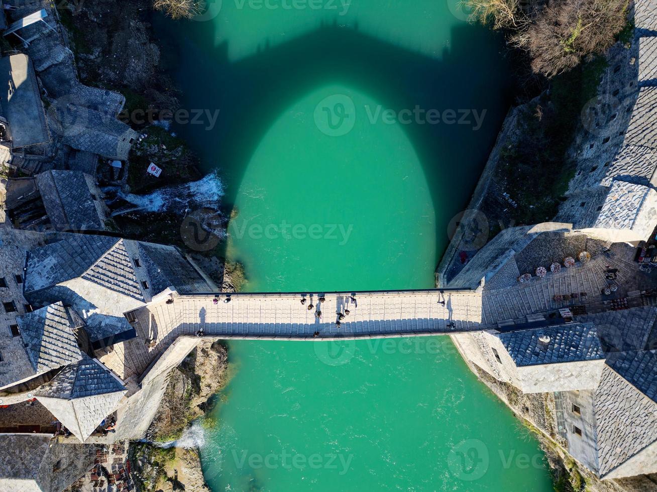 Aerial drone view of the Old Bridge in Mostar city in Bosnia and Herzegovina during sunny day. Blue turquoise colors of Neretva river. Unesco World Heritage Site. People walking over the bridge. photo