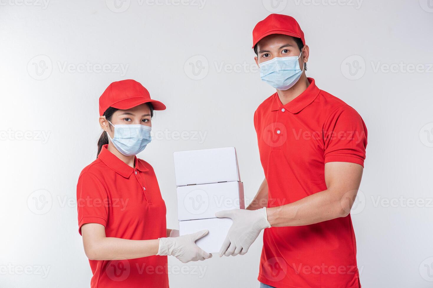 Image of a conscious young delivery man in red cap blank t-shirt uniform face mask gloves standing with empty white cardboard box isolated on light gray background studio photo