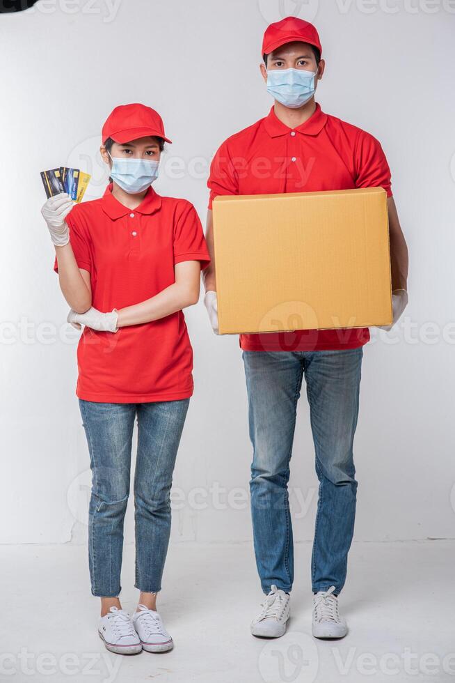 Image of a conscious young delivery man with credit card in red cap blank t-shirt uniform face mask gloves standing with empty brown cardboard box isolated on light gray background studio photo