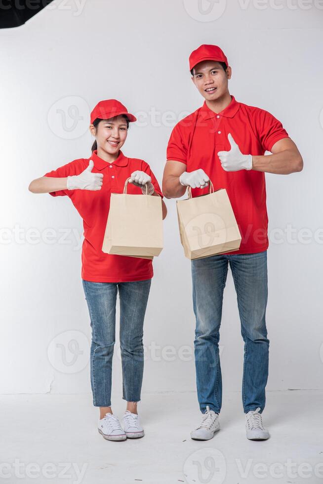 imagen de un joven repartidor feliz con gorra roja en blanco uniforme de camiseta de pie con un paquete de papel artesanal marrón vacío aislado en un estudio de fondo gris claro foto
