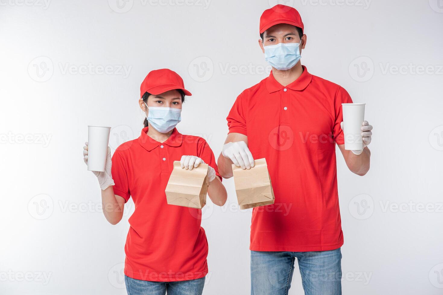 Image of a happy young delivery man in red cap blank t-shirt uniform face mask gloves standing with empty brown craft paper packet isolated on light gray background studio photo