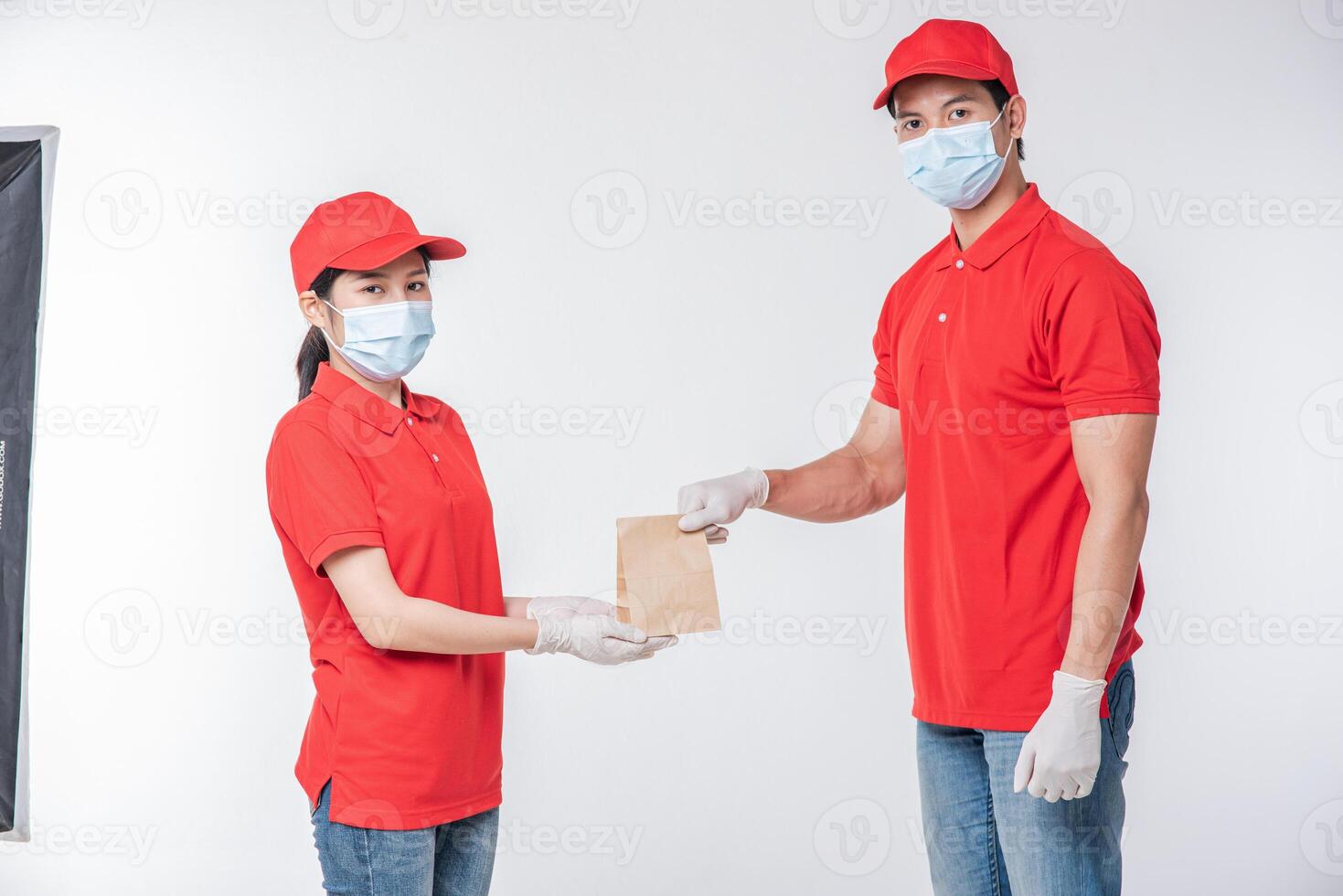 imagen de un joven repartidor feliz con gorra roja en blanco camiseta uniforme mascarilla guantes de pie con un paquete de papel artesanal marrón vacío aislado en un estudio de fondo gris claro foto