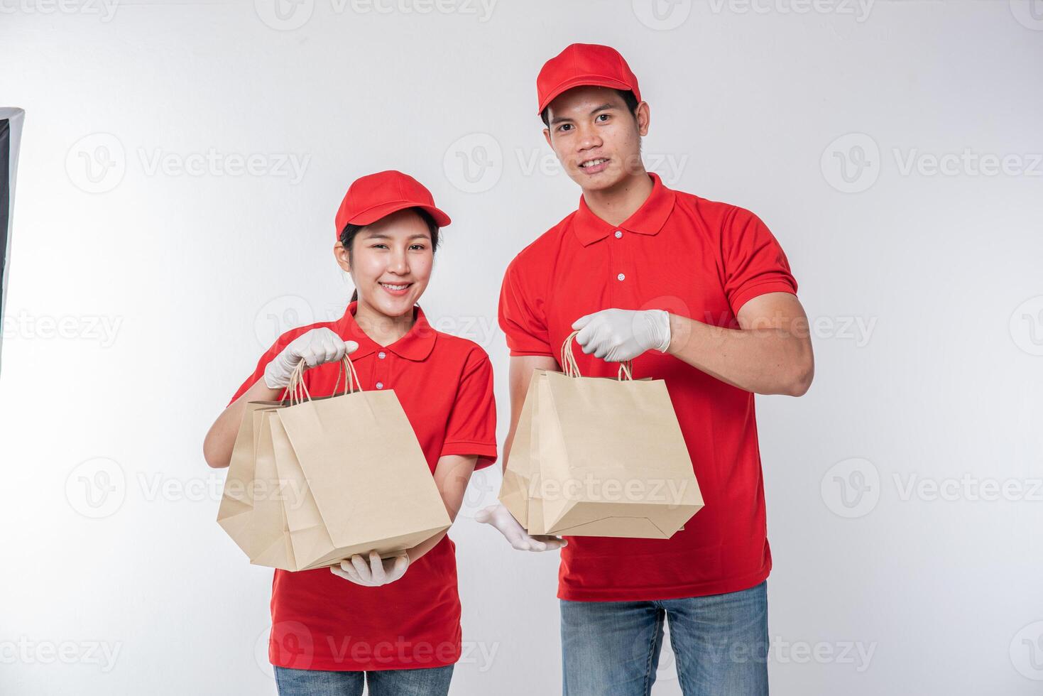 Image of a happy young delivery man in red cap blank t-shirt uniform standing with empty brown craft paper packet isolated on light gray background studio photo