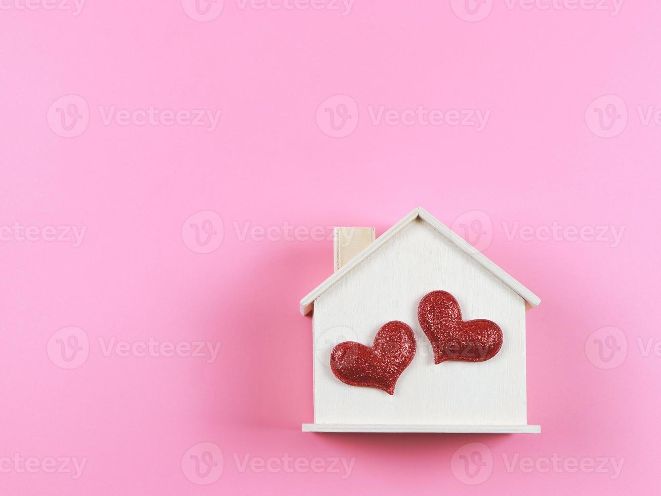 endecha plana de casa modelo de madera con dos corazones rojos brillantes sobre fondo rosa. casa de ensueño, hogar de amor, relación fuerte, san valentín. foto