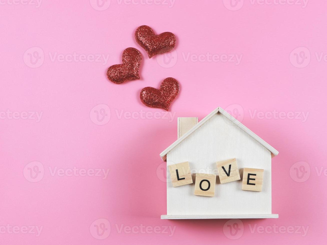 flat lay of wooden model house with wooden letter L O V E on it and red glitter hearts on pink  background. dream house , home of love, strong relationship, valentines. photo