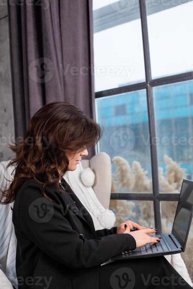 Beautiful businesswoman working on a laptop in a chair near the window. Female freelancer connecting to internet via computer. Blogger or journalist writing new article. photo