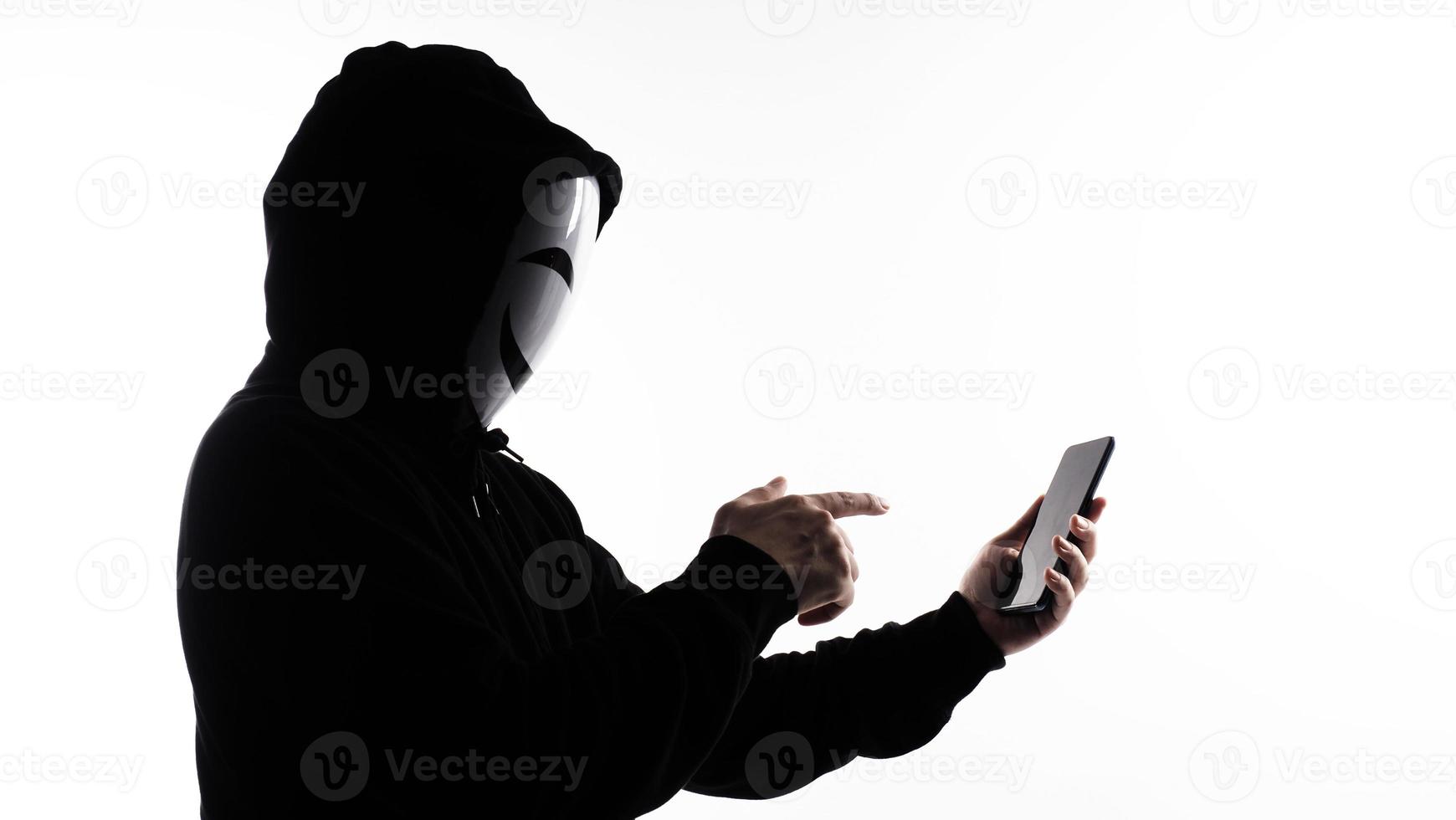 Hacker Anonymous and face mask with smartphone in hand. Man in black hood shirt holding and using mobile phone on white background. Represent cyber crime data hacking or stealing personal data concept photo