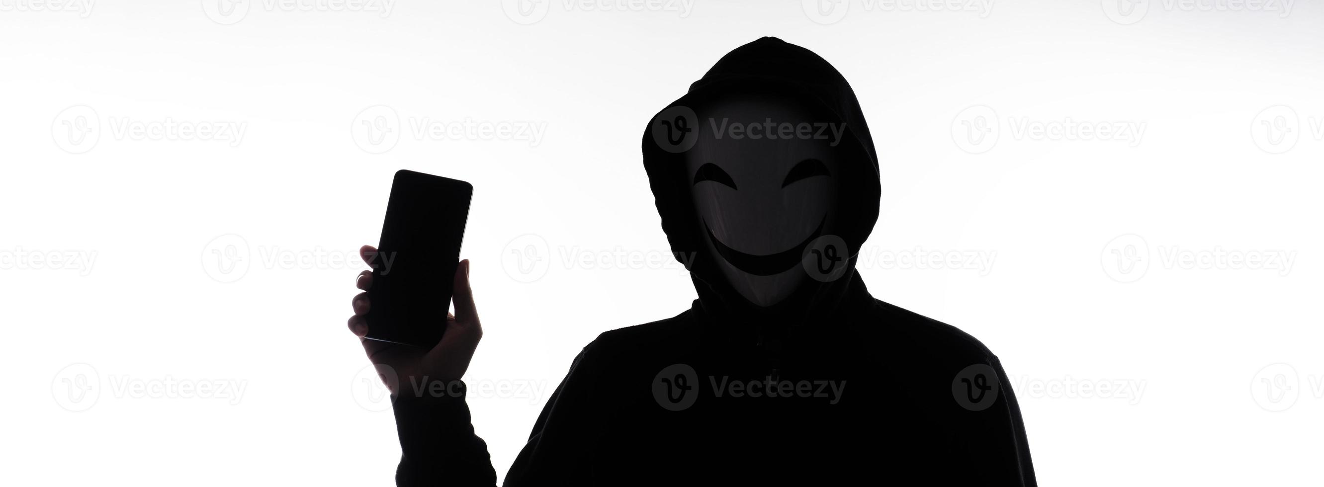 hacker anónimo y mascarilla con smartphone en la mano. hombre con camisa de capucha negra sosteniendo y usando un teléfono móvil con fondo blanco. representan el concepto de piratería informática o robo de datos personales foto