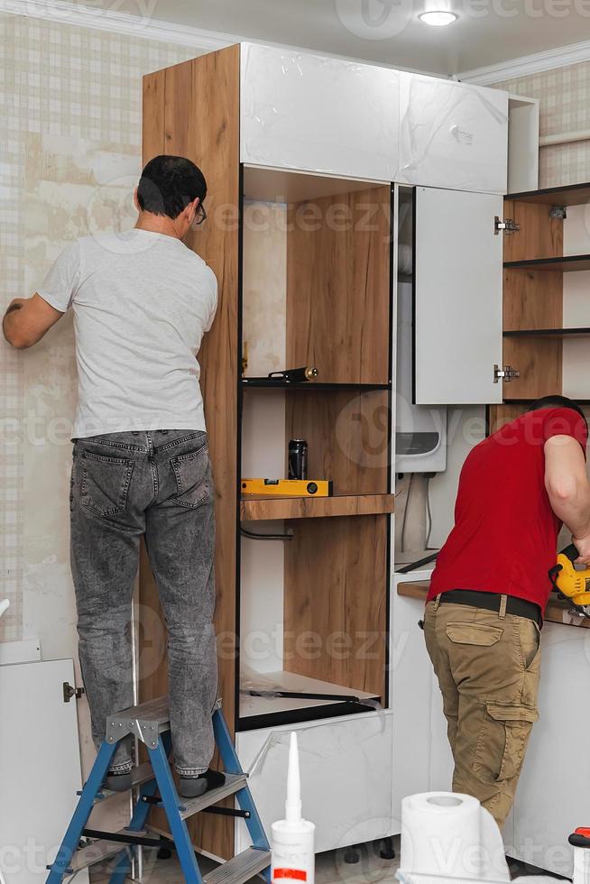 Two craftsmen, furniture makers and assemblers, are installing a modern kitchen in a home, tools in their hands. Concept furniture to order, improvement of living conditions, repair photo