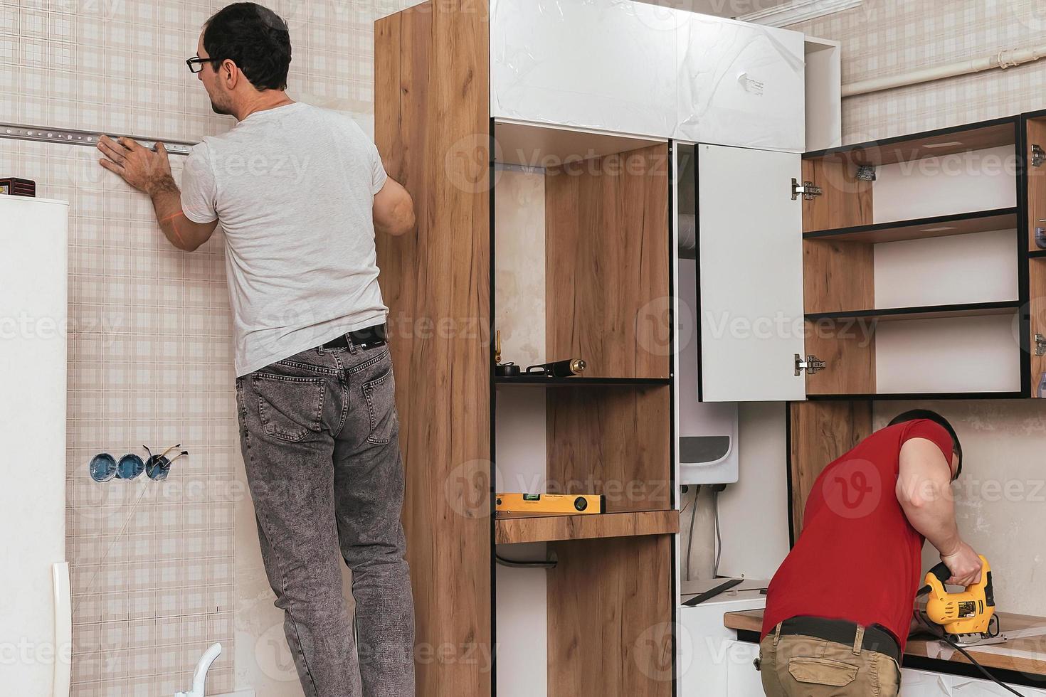 Two craftsmen, furniture makers and assemblers, are installing a modern kitchen in a home, tools in their hands. Concept furniture to order, improvement of living conditions, repair photo
