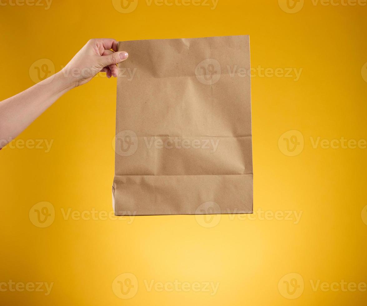 female holding in hand brown blank craft paper bag for takeaway on yellow background. Packaging template mock up. Delivery service concept photo