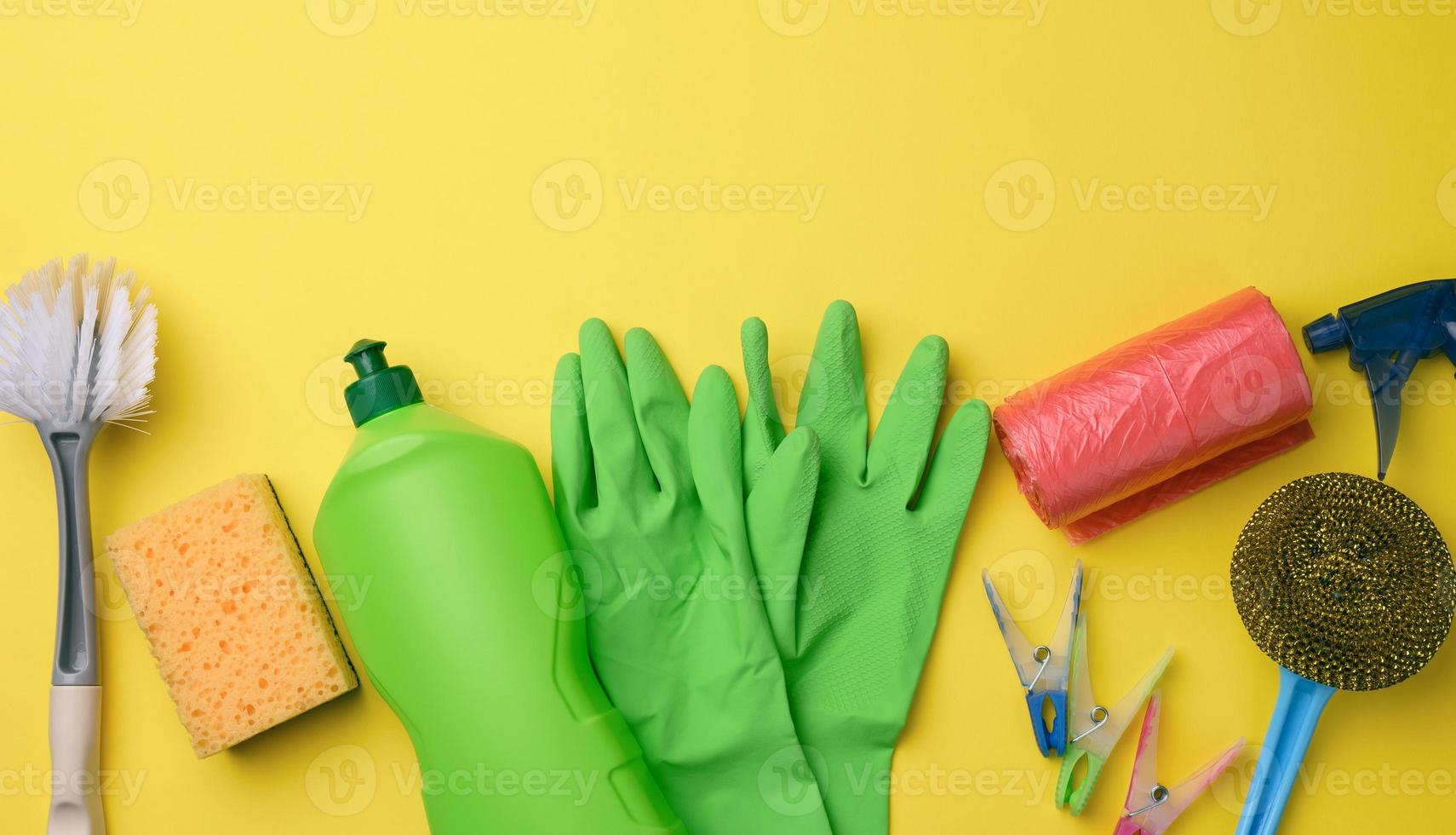 rubber green gloves for cleaning, red trash can plastic bag roll and plastic bottle with detergent, brush on yellow background photo
