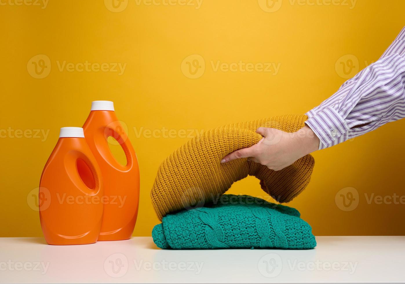 washed folded clothes and plastic orange large bottles with liquid detergent stand on a white table, yellow background. Routine homework photo