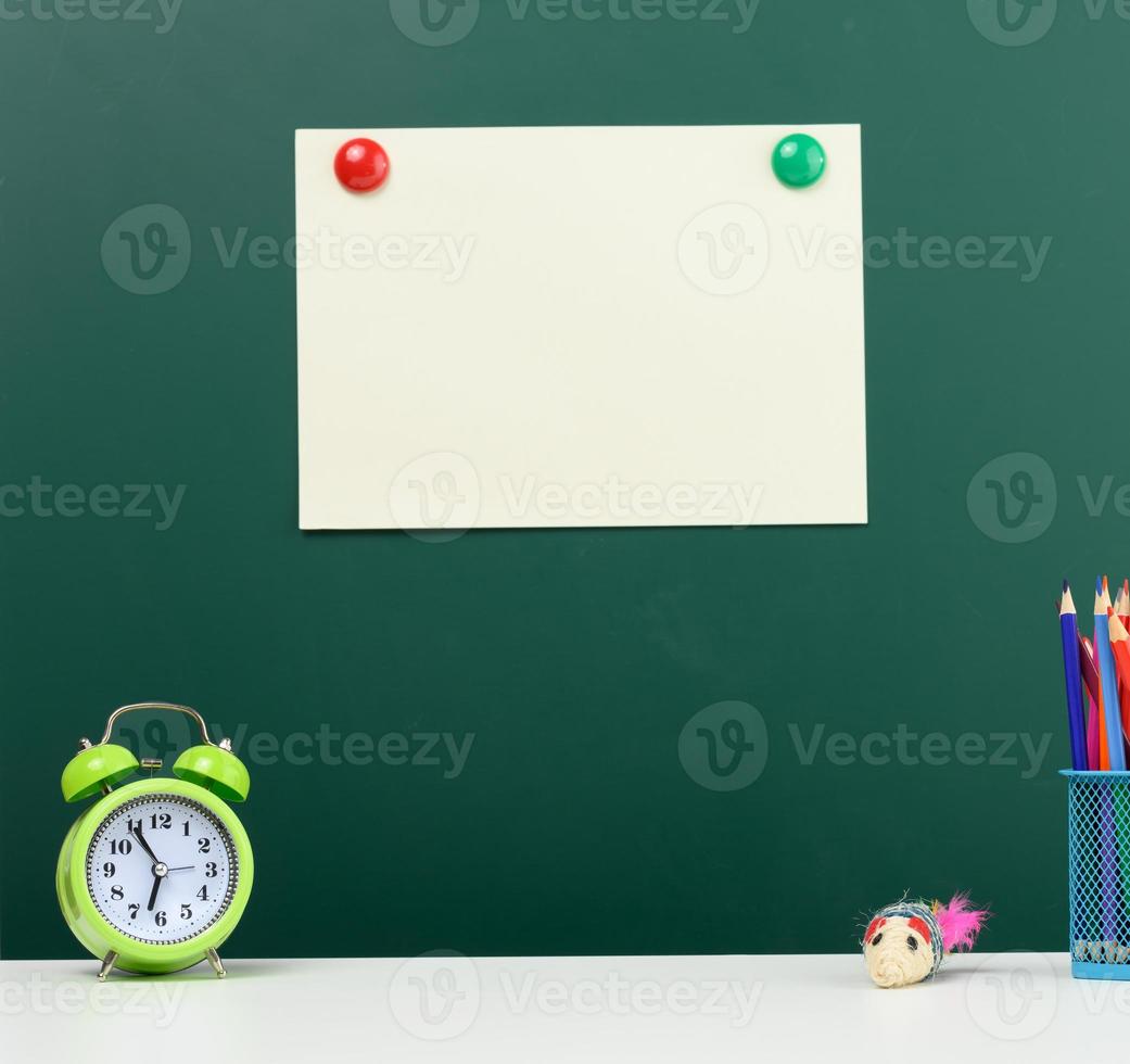 blank sheet of paper affixed on a green chalk board, alarm clock and multi-colored pencils. Back to school photo