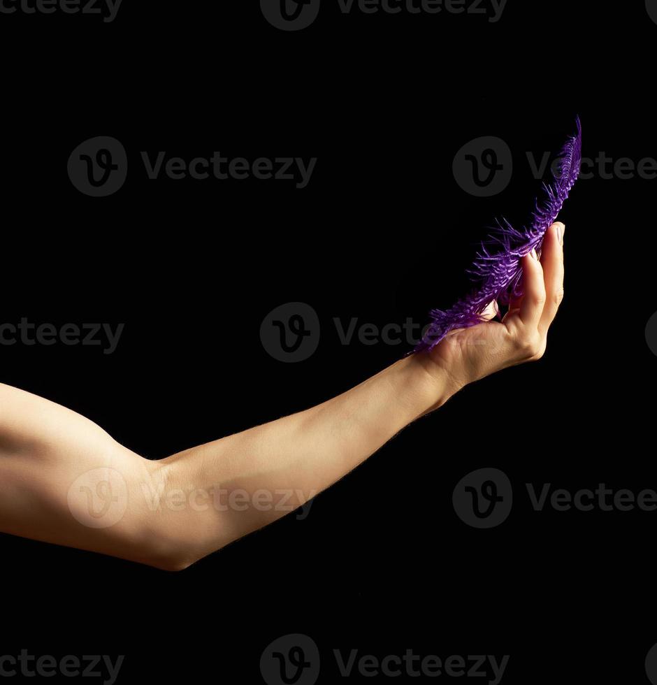 female hand is bent at the elbow, the muscles are tense and hold a blue feather on a dark background photo