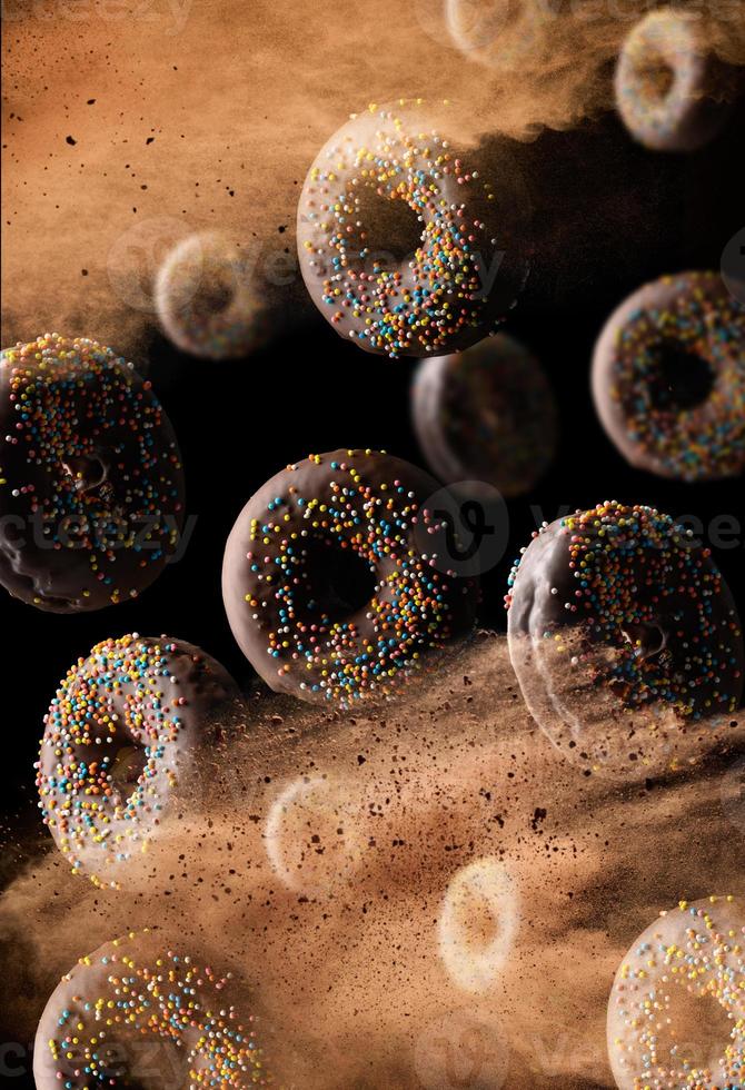 chocolate round donuts with multicolored sugar sprinkles levitate in a cloud of brown cocoa on a black background. Powder flies up photo