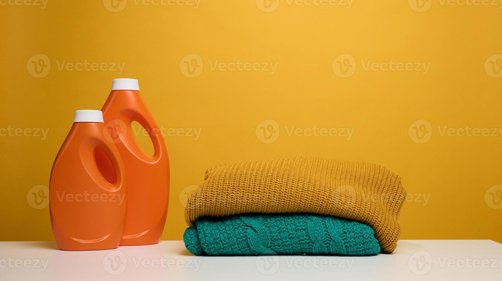 pila de ropa doblada lavada y botella grande de plástico naranja con soporte de detergente líquido sobre una mesa blanca, fondo amarillo. tarea de rutina foto