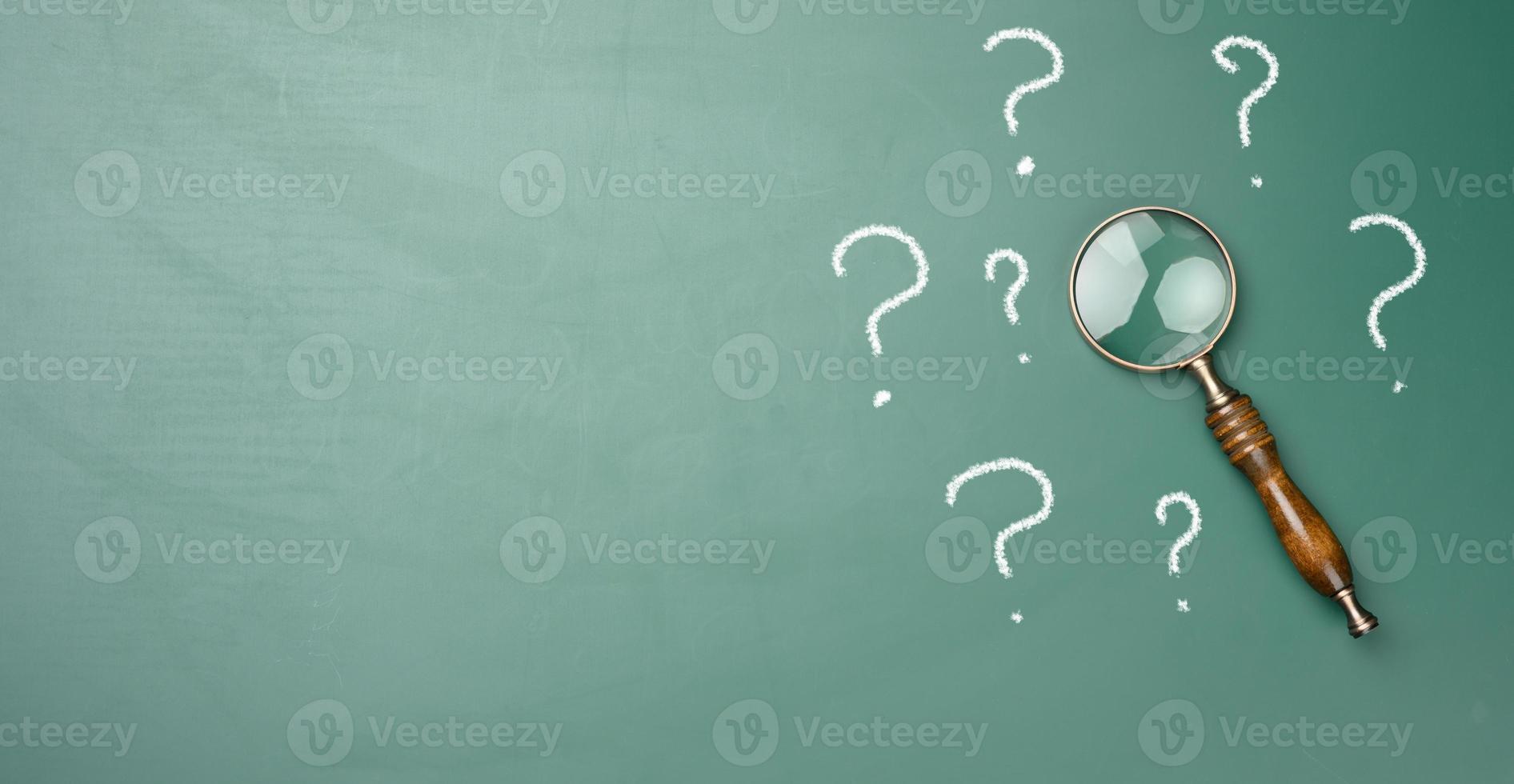 wooden magnifying glass and white chalk drawn question marks on a green chalk board. The concept of finding solutions, true or false photo