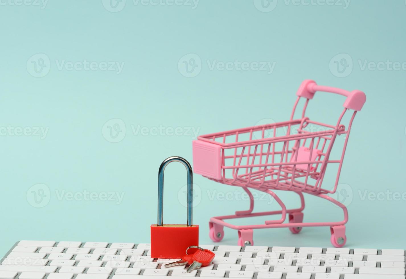 a red metal padlock stands on the keyboard and behind a miniature shopping cart, blue background photo