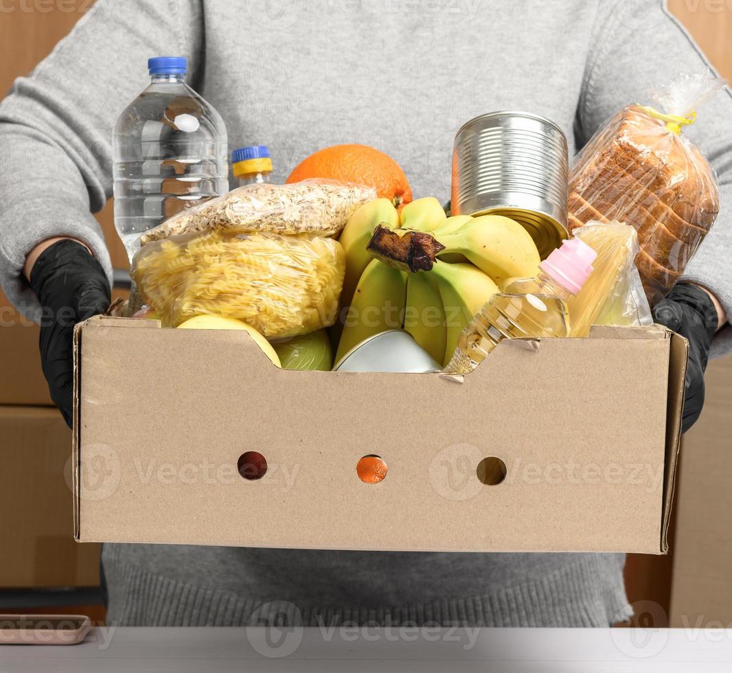 mujer con suéter gris y guantes negros sosteniendo una caja de cartón con comestibles, concepto de asistencia y voluntariado foto