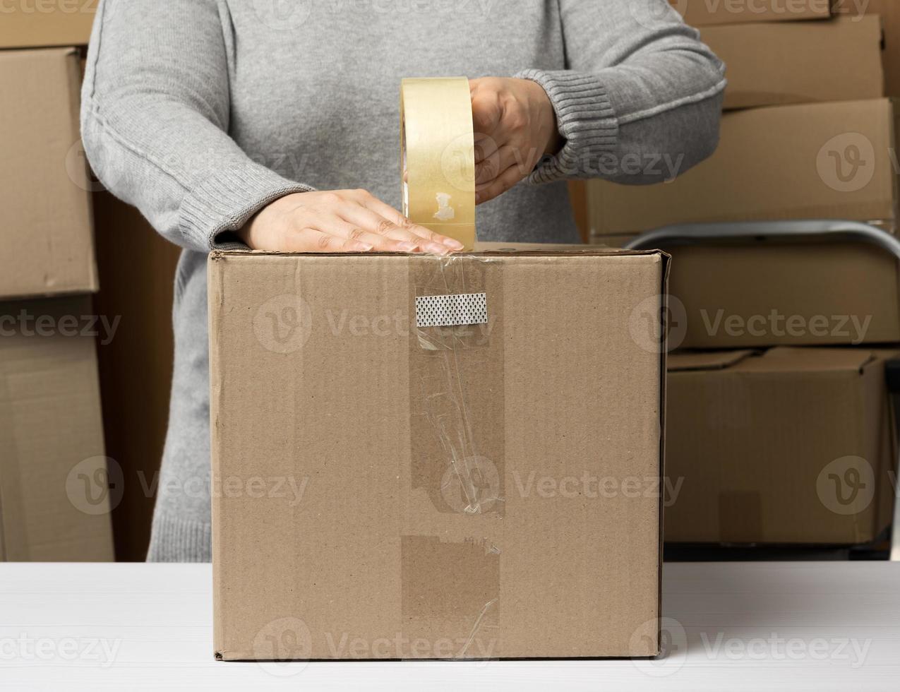 woman in a gray sweater holds a roll of duct tape and packs brown cardboard boxes on a white table photo