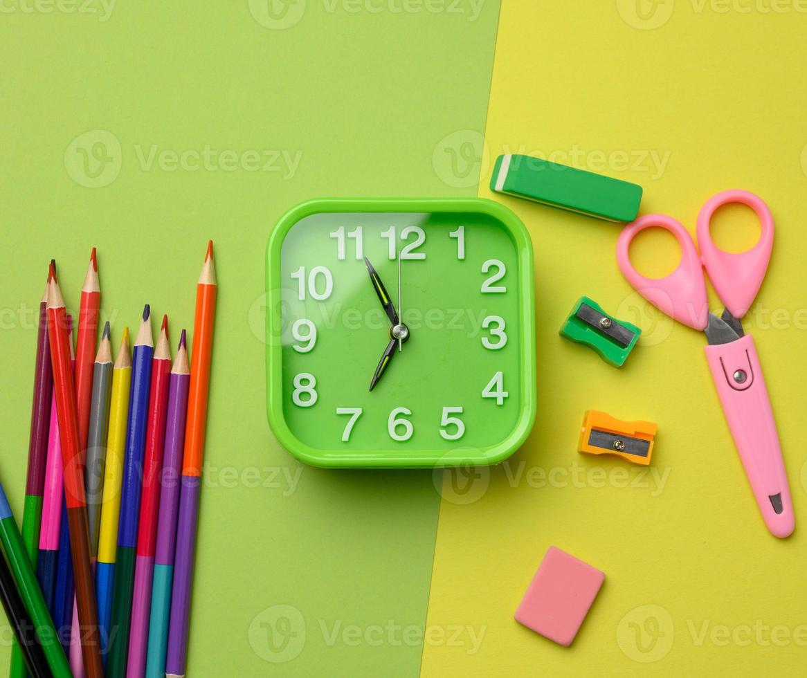 square green clock and stack of multicolored wooden pencils on a green background, top view photo