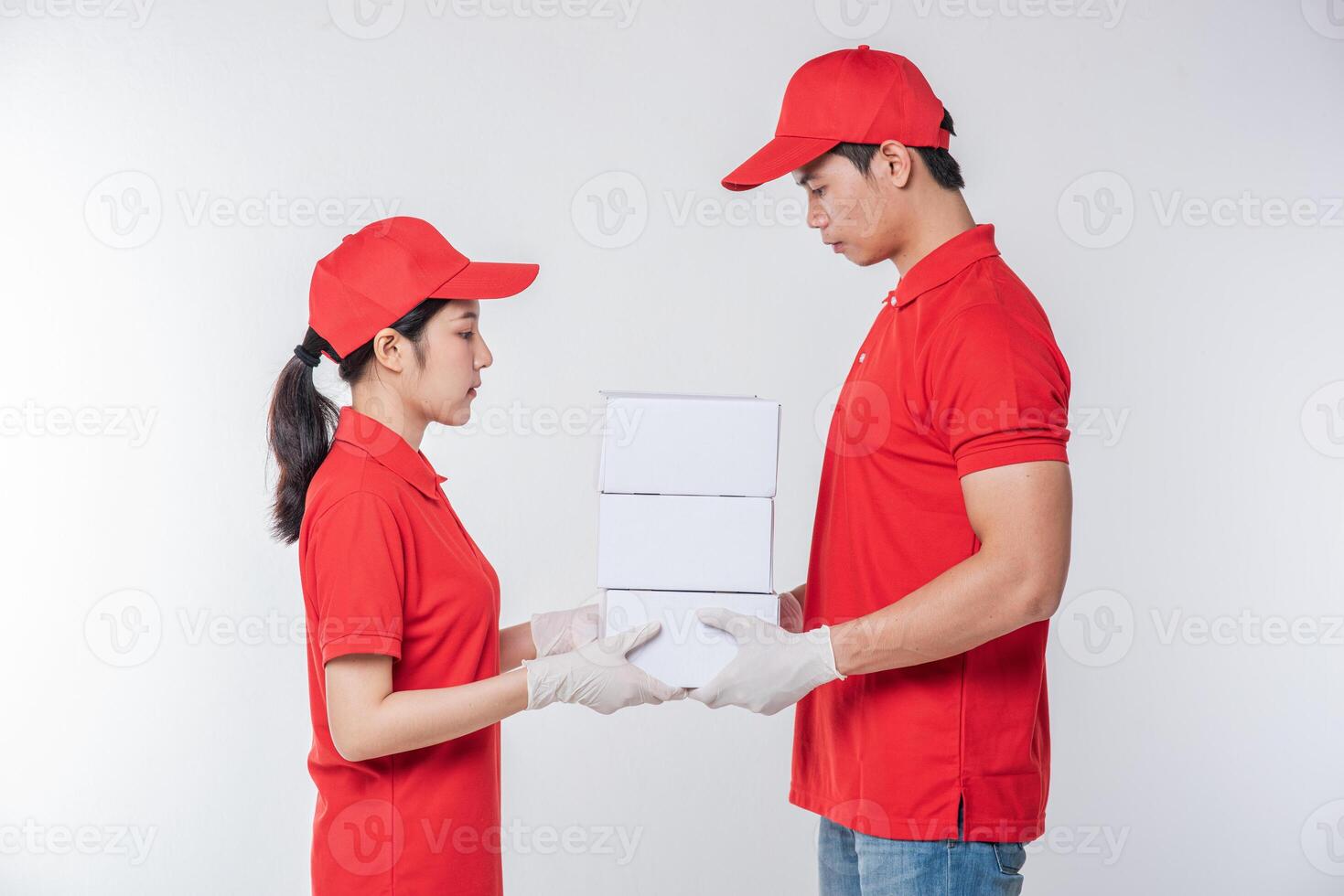 imagen de un joven repartidor feliz con gorra roja en blanco uniforme de camiseta de pie con una caja de cartón blanca vacía aislada en un estudio de fondo gris claro foto