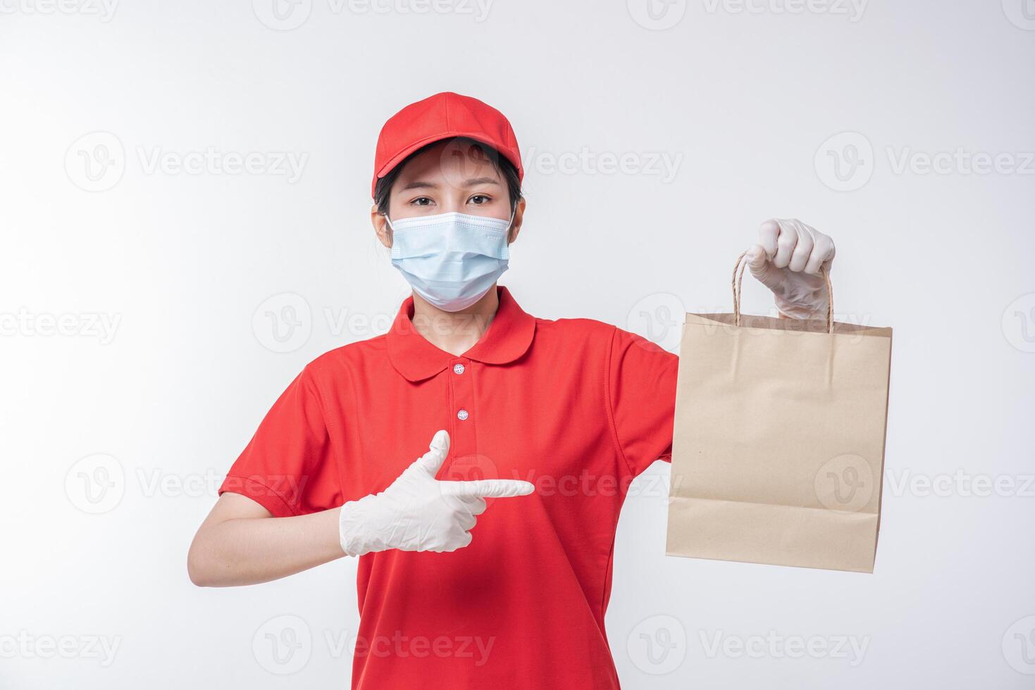 imagen de un joven repartidor feliz con gorra roja en blanco camiseta uniforme mascarilla guantes de pie con un paquete de papel artesanal marrón vacío aislado en un estudio de fondo gris claro foto