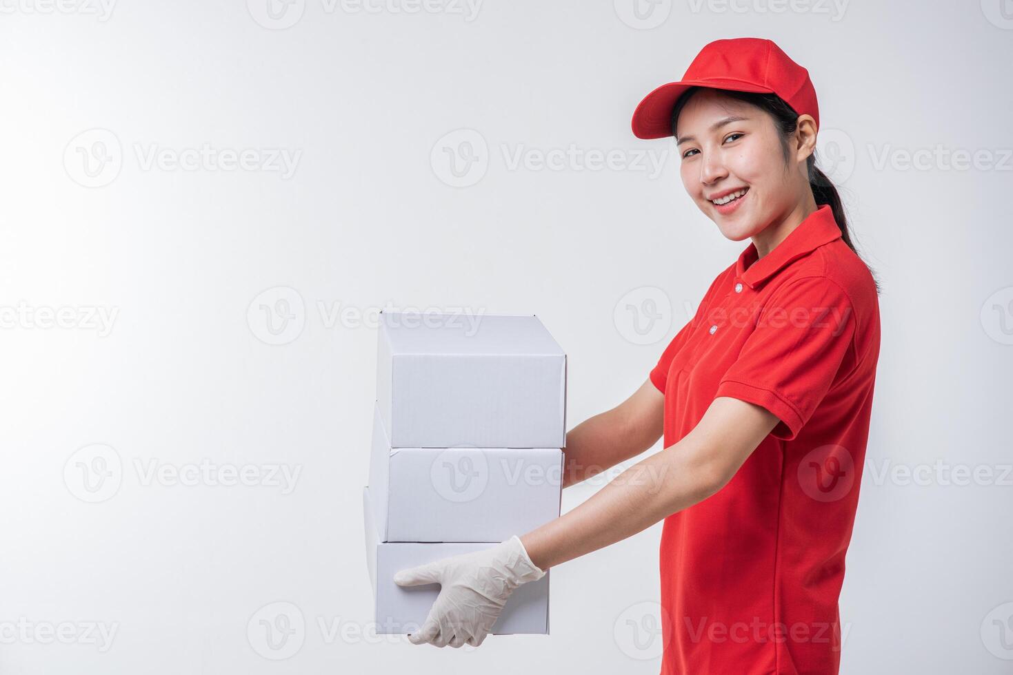 imagen de un joven repartidor con uniforme de camiseta en blanco con gorra roja de pie con una caja de cartón blanca vacía aislada en un estudio de fondo gris claro foto