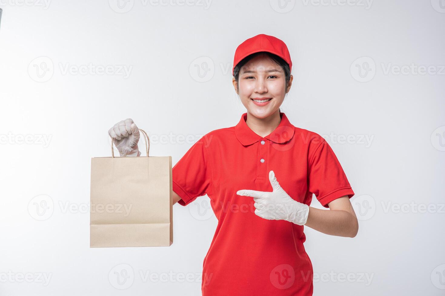 imagen de un joven repartidor feliz con gorra roja en blanco uniforme de camiseta de pie con un paquete de papel artesanal marrón vacío aislado en un estudio de fondo gris claro foto