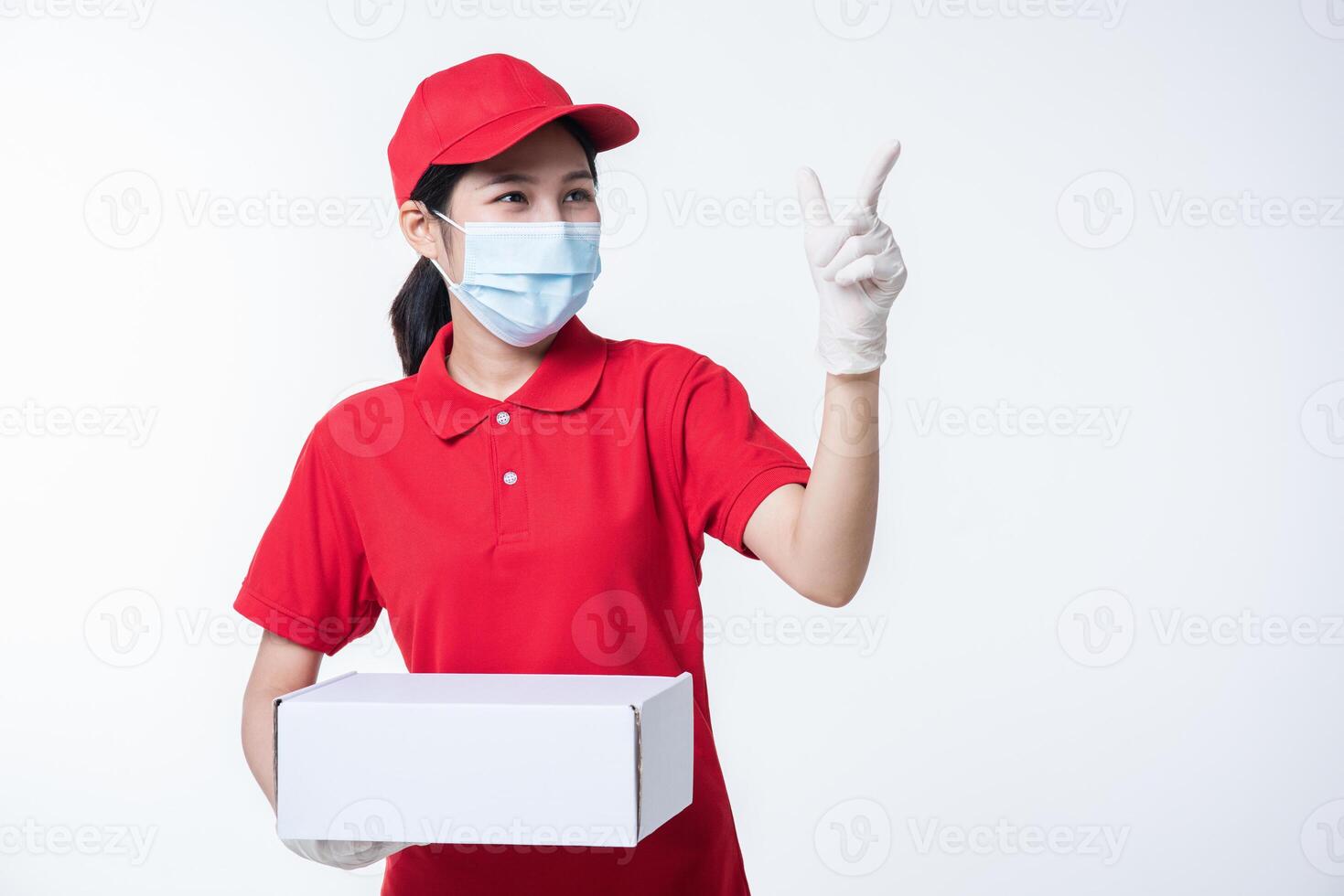 imagen de un joven repartidor consciente con gorra roja en blanco, camiseta uniforme, guantes de máscara facial de pie con una caja de cartón blanca vacía aislada en un estudio de fondo gris claro foto