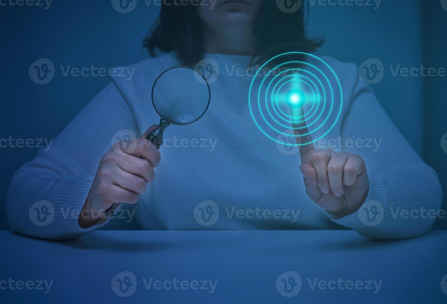 a woman sits at a table and holds a magnifying glass, with the other hand presses on a virtual screen with neon circles. Search for information, start the program photo