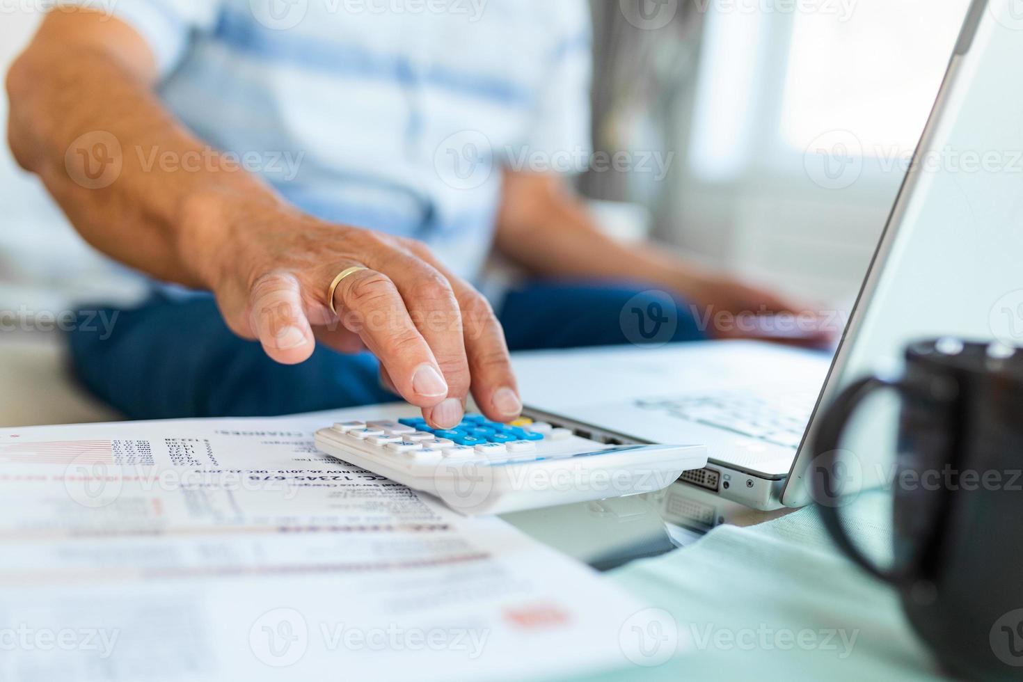 Serious elder senior grey-haired grandpa using calculator counting bank loan payment holding papers at home, focused mature old man doing paperwork calculating bills managing finances sit on sofa photo