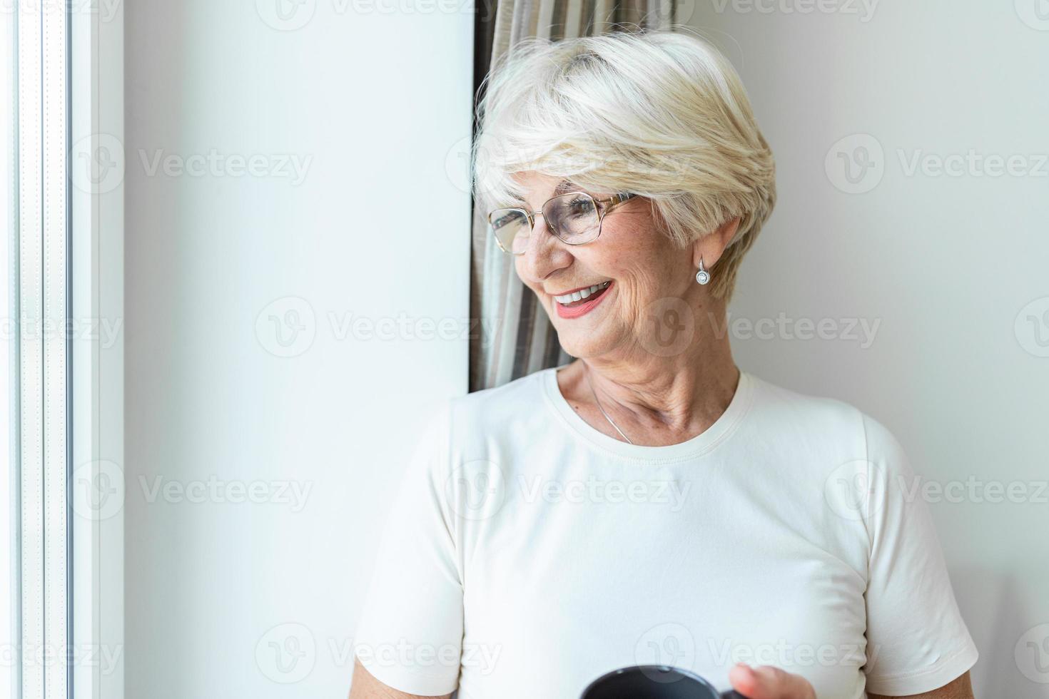 Happy senior woman day dreaming while looking through the window and enjoying in cup of coffee. Positive mature woman in white cotton blouse drinking mug of coffee and looking through window photo