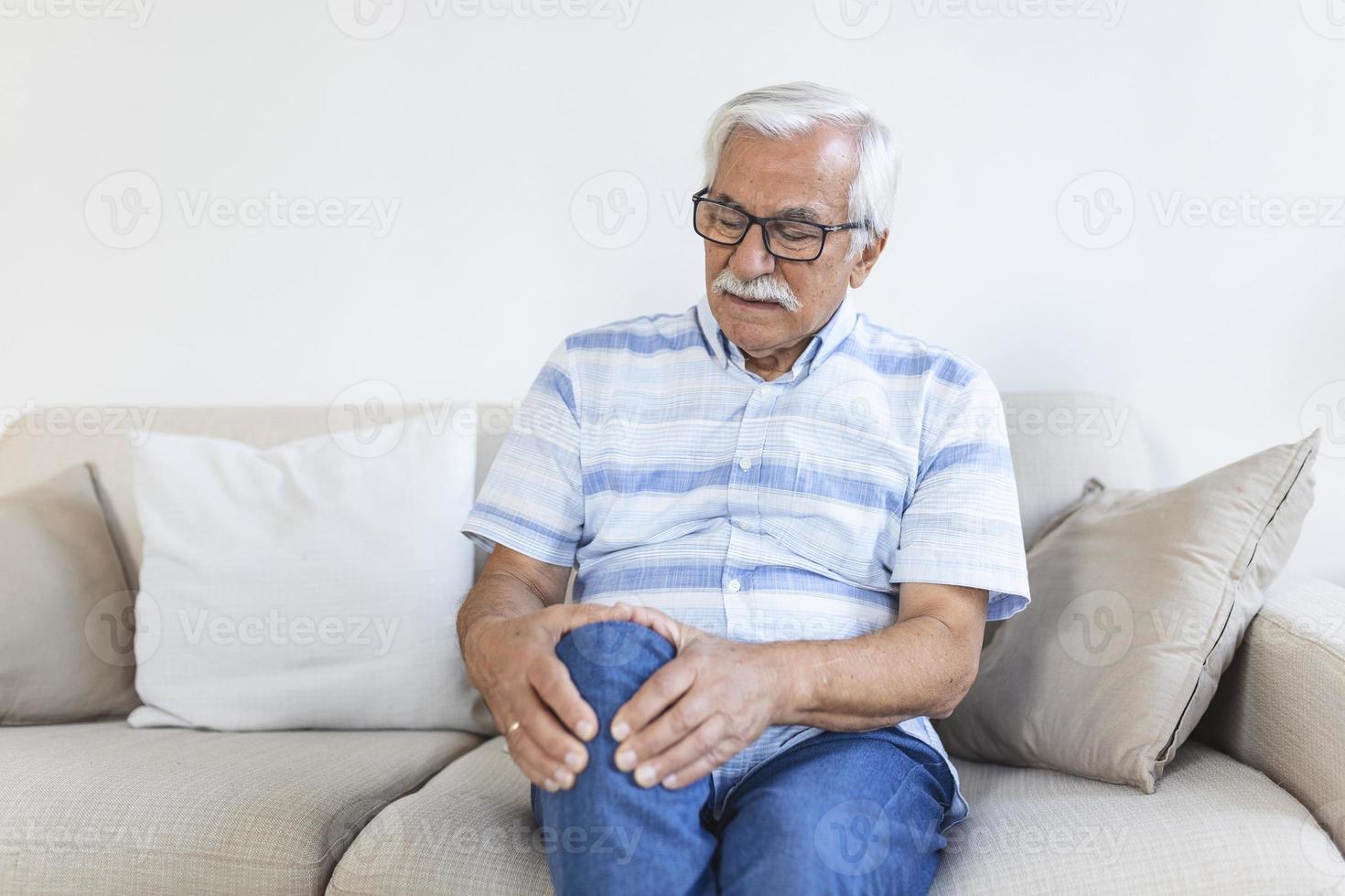 Elderly man sitting on a sofa at home and touching his painful knee. people, health care and problem concept - unhappy senior man suffering from knee ache at home photo