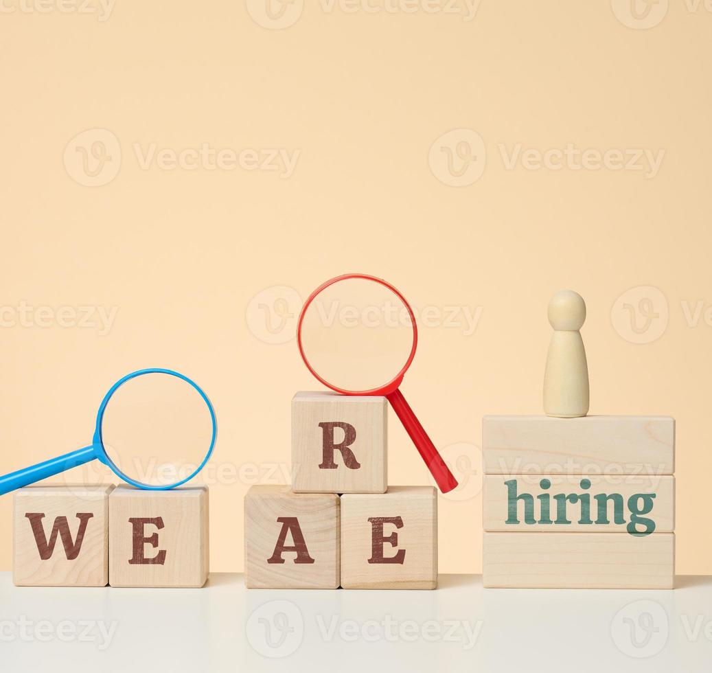 the inscription we are hiring and a wooden figurine on the podium with a plastic magnifying glass on a beige background. Employee search concept photo