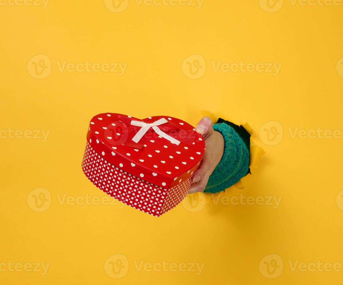 hembra mano sostiene un rojo caja con un regalo en un amarillo fondo, parte de el cuerpo palos fuera de un Rasgado agujero en un papel antecedentes. felicidades, fiesta sorpresa foto