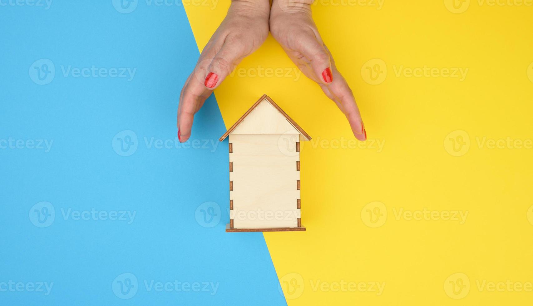 two female hands folded to each other over a wooden miniature model house on a yellow-blue background. Real estate insurance concept photo