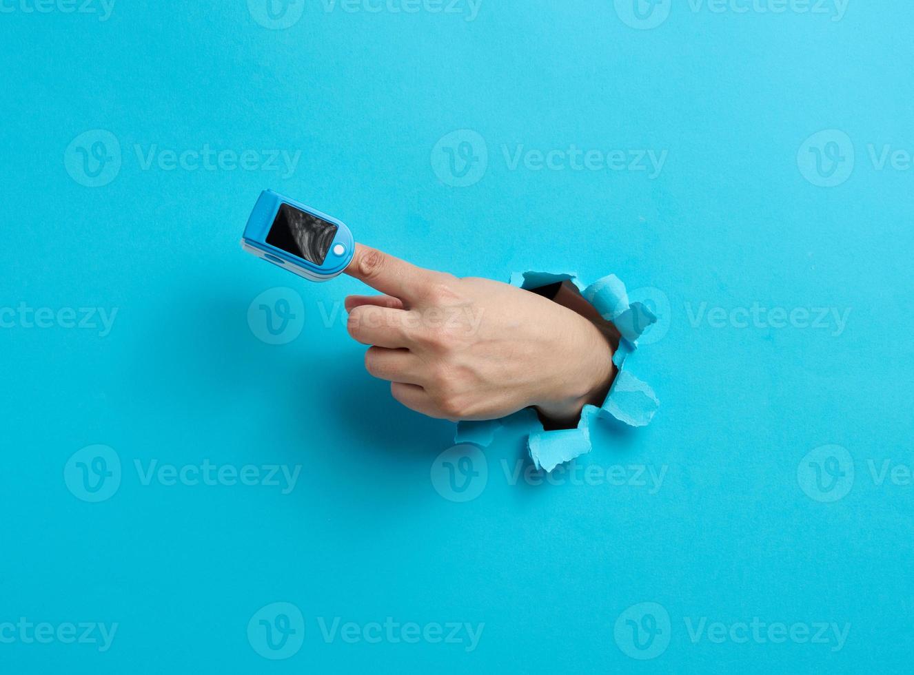 an oximeter for measuring oxygen in the blood is worn on the index finger, part of the hand is sticking out of a torn hole in the blue paper photo