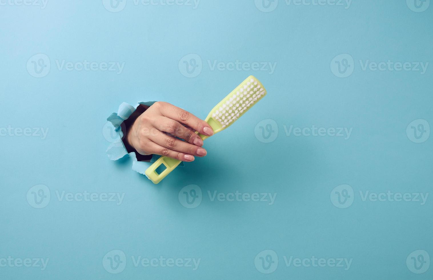 female hand holds a green plastic brush on a blue background. A part of the body sticks out of a hole with torn edges in a paper background photo