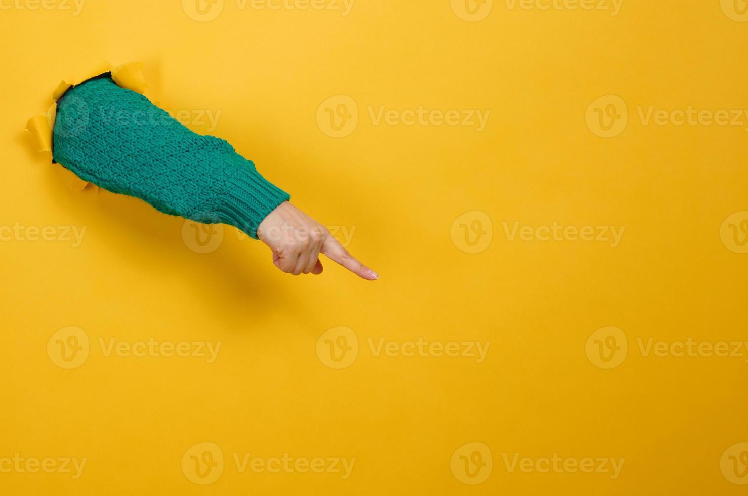 female hand sticks out of a torn hole in a yellow paper background, part of the body points with the index finger to the side. Copy space photo