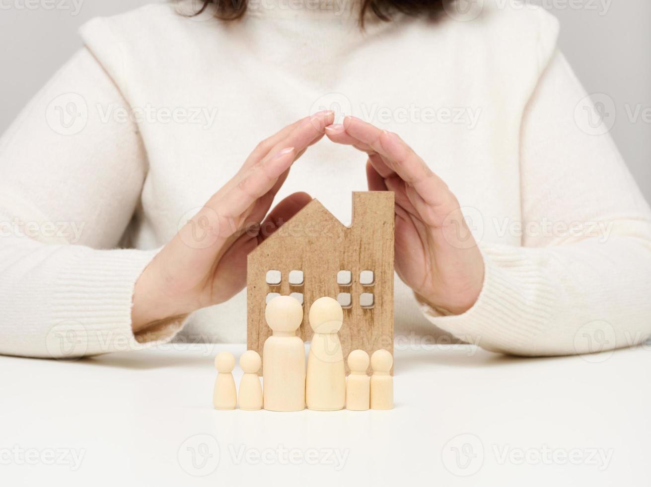 a wooden house and figures of a family, two female palms protect objects from above. Real estate insurance concept, happy family photo