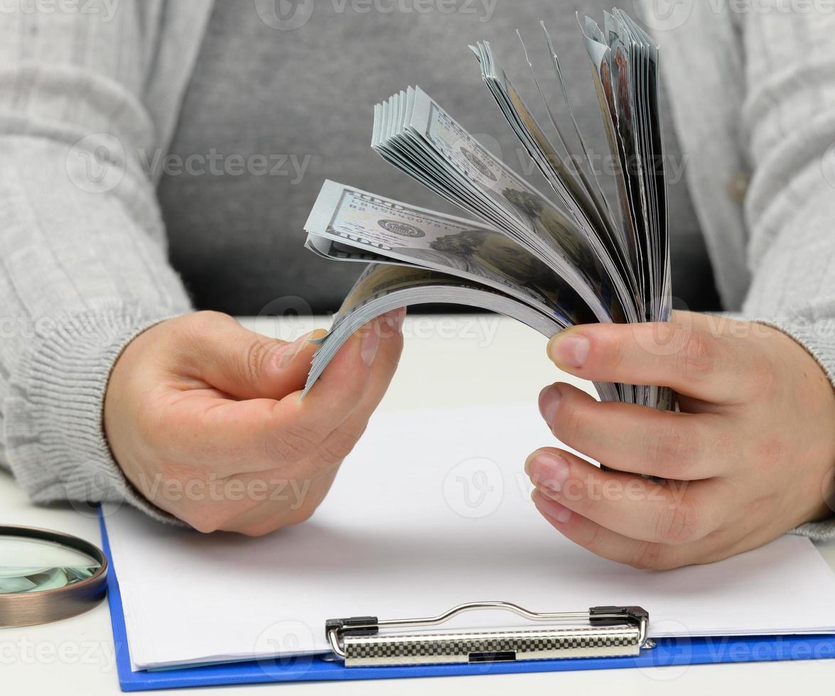 a stack of one hundred dollar bills in a woman's hand and a wooden magnifying glass on a white table. Budget analysis, income and expenses, profit calculation photo